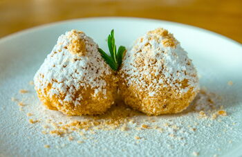 Zwei Quarkknödel, ummantelt mit gemahlenen Haselnüssen und Puderzucker, auf einem weißen Teller. Dazwischen ein kleines Minzblatt.