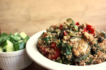 Ein Couscous-Salat in einer großen weißen Schüssel mit geschnittenem Gemüse und frischen Kräutern. Links daneben eine kleine weiße Schüssel mit gewürfelten Gurken.