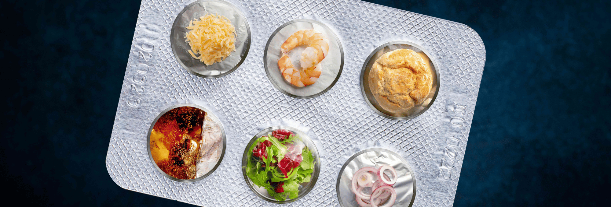 Das Bild zeigt einen Blister, in dem statt Tabletten kleine Lebensmittel abgebildet sind. Z.B. ein Brötchen, 2 Shrimps und einige Zwiebeln.