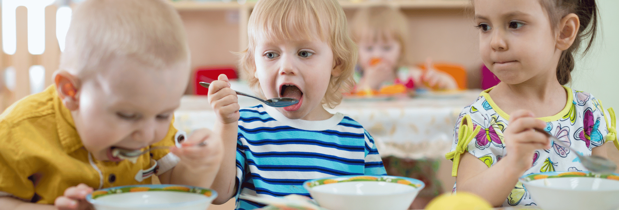 Das Bild zeigt drei kleine Kinder an einem Kitatisch, die gemeinsam essen.